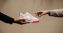 Pink and white sneaker being passed between two hands against a beige background
